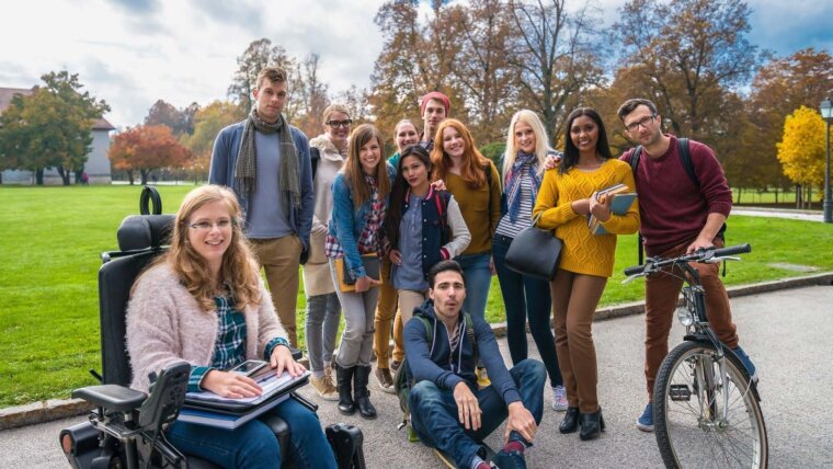 The photo shows a group of students on campus in front of a lawn, including men and women of different skin colors and a female student in a wheelchair.