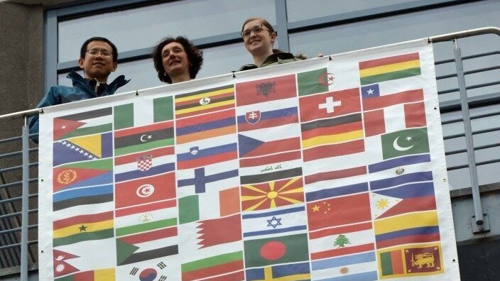 Vizepräsidentin für Studium und Lehre präsentiert mit zwei Studierenden das Banner "Denken ohne Grenzen" der Universität mit den Flaggen vieler Länder