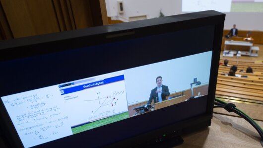 Ein Monitor im Vordergrund zeigt die digitale Version einer Lehrveranstaltung, die im Hintergrund im Hörsaal gehalten wird.