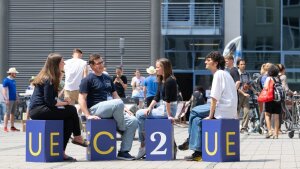 Auf dem Campus Ernst-Abbe-Platz sitzen junge Menschen auf blauen Hockern, die zusammen "EC2U" buchstabieren.