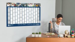 Neben einer am Schreibtisch sitzenden Frau ist der Wandkalender 2024 der Uni Jena zu sehen.