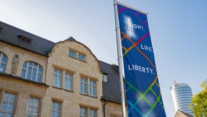 A flag with the slogan "Light, Life, Liberty" in front of the main university building.