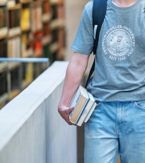 Ein STudent trägt einen Stapel Bücher in der Bibliothek